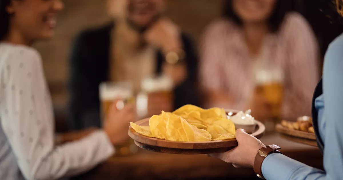 Garçom servindo prato de batata chips em tábua de madeira para grupo de clientes em lanchonete, com cervejas na mesa e pessoas conversando ao fundo