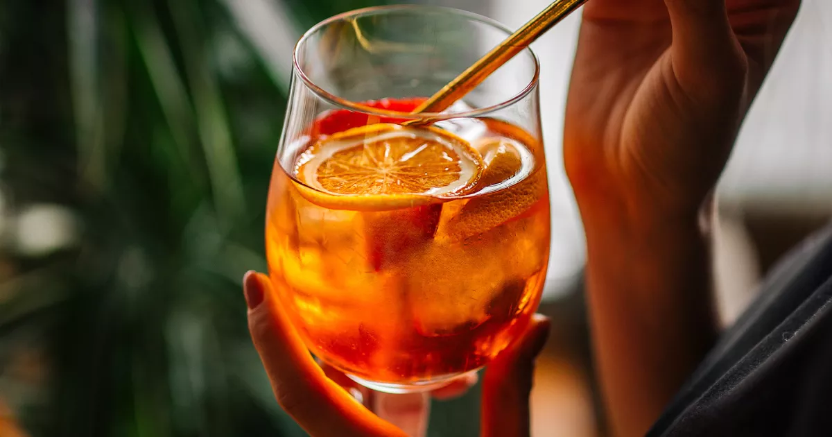 Mão segurando taça de vinho com Aperol Spritz laranja, fatia de laranja desidratada e colher dourada, planta verde desfocada ao fundo