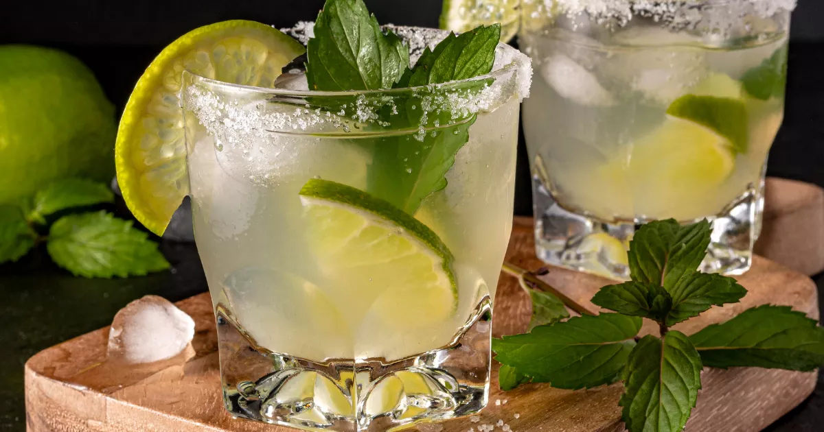 Dois copos rocks com Caipirinha e borda de sal sobre tábuas de madeira, decorados com fatias de limão e folhas de hortelã frescas