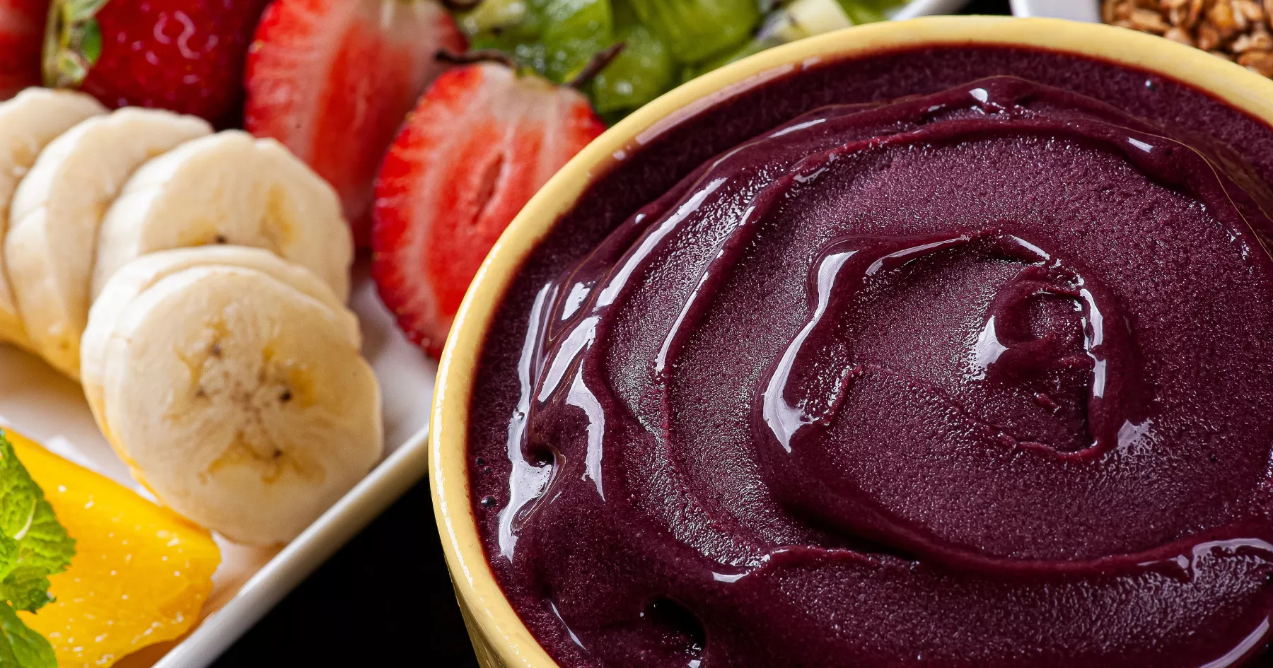 Creme de açaí com textura firme e brilhante em tigela, ao lado de banana, morango, uva, laranja e granola porcionados em prato branco para a montagem