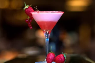 Cosmopolitan rosa em taça de martini com borda de açúcar, espuma no topo e guarnição de morango e groselha vermelha, fundo de bar desfocado