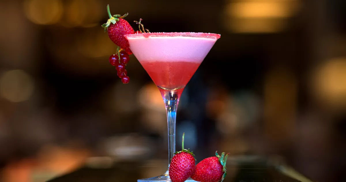 Cosmopolitan rosa em taça de martini com borda de açúcar, espuma no topo e guarnição de morango e groselha vermelha, fundo de bar desfocado
