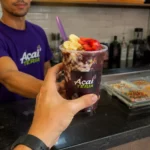 Atendente de açaiteria de camiseta roxa entrega copo de açaí com banana, morango e granola ao cliente no balcão, com potes de complementos e cardápio de preços ao fundo