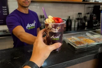 Atendente de açaiteria de camiseta roxa entrega copo de açaí com banana, morango e granola ao cliente no balcão, com potes de complementos e cardápio de preços ao fundo