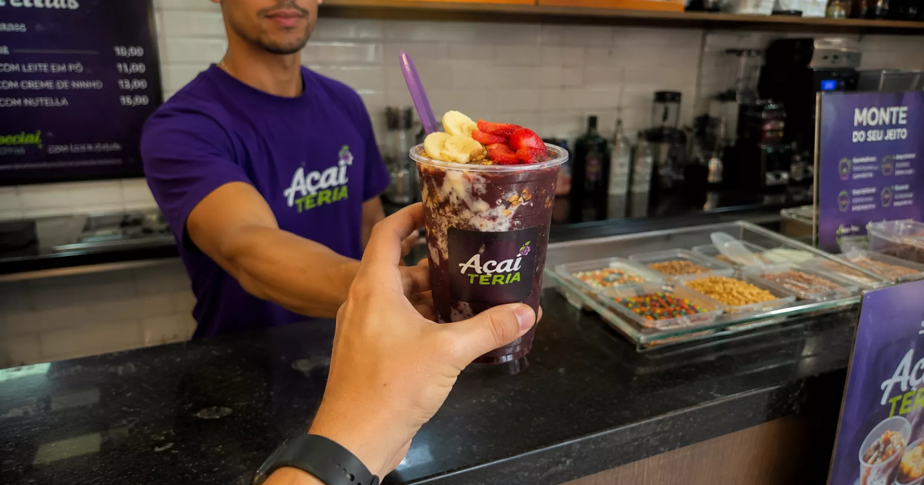 Atendente de açaiteria de camiseta roxa entrega copo de açaí com banana, morango e granola ao cliente no balcão, com potes de complementos e cardápio de preços ao fundo