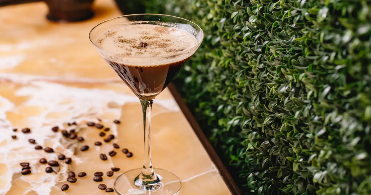 Espresso Martini em taça de martini com espuma de café cremosa e grão de café sobre mesa de ladrilhos com grãos espalhados, muro de plantas ao fundo
