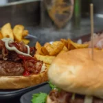 Dois hambúrgueres artesanais em bandejas escuras com batata rústica, um deles aberto mostrando o blend com bacon, cebola e maionese, balcão de hamburgueria ao fundo