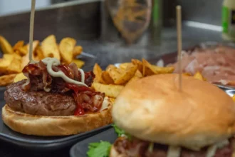 Dois hambúrgueres artesanais em bandejas escuras com batata rústica, um deles aberto mostrando o blend com bacon, cebola e maionese, balcão de hamburgueria ao fundo