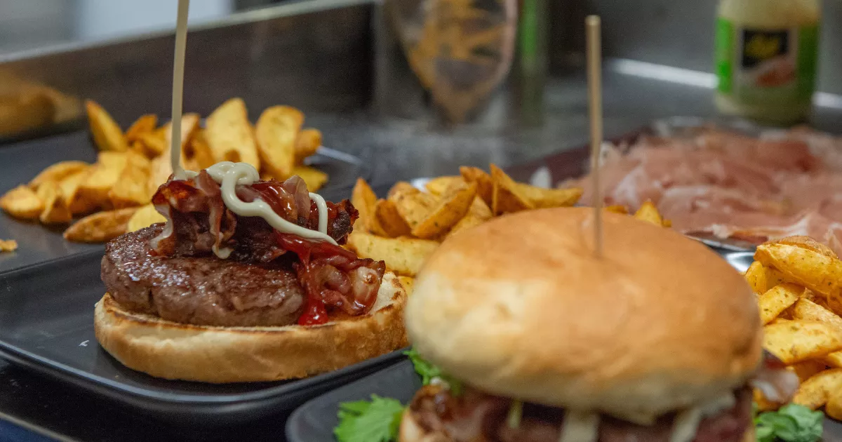 Dois hambúrgueres artesanais em bandejas escuras com batata rústica, um deles aberto mostrando o blend com bacon, cebola e maionese, balcão de hamburgueria ao fundo