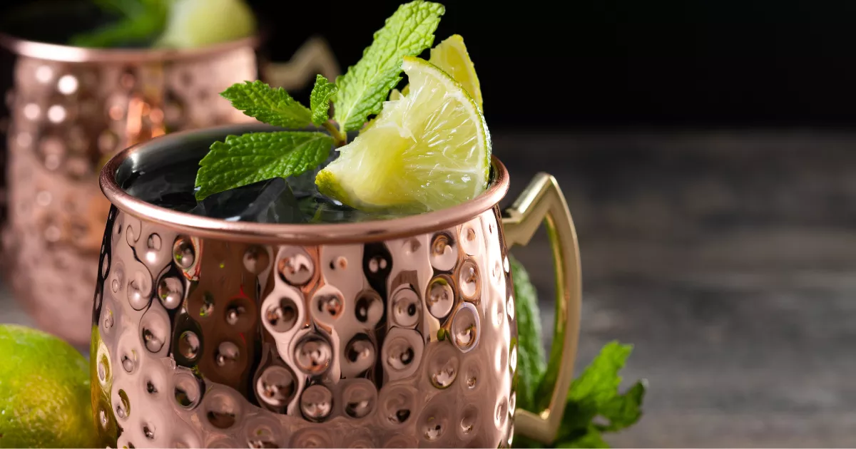 Caneca de cobre martelada com Moscow Mule decorada com cunha de limão e folhas de hortelã frescas, segunda caneca visível ao fundo escuro