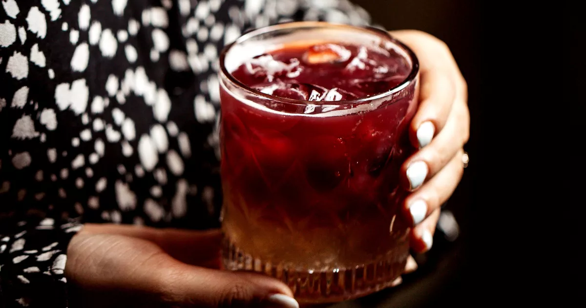 Duas mãos segurando copo rocks com Negroni vermelho escuro e cubos grandes de gelo, blusa preta e branca estampada ao fundo