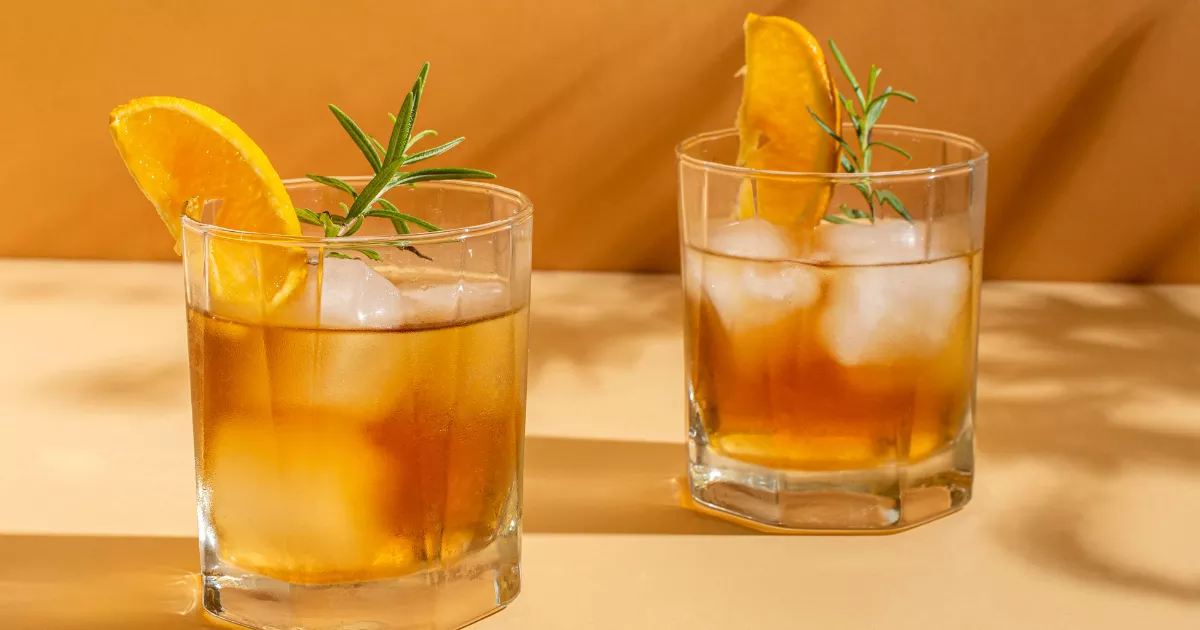 Dois copos old fashioned com Whisky Sour âmbar decorados com fatia de laranja e ramo de alecrim, sobre superfície dourada com luz quente