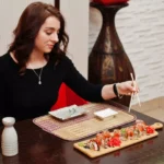 Mulher sentada em mesa de restaurante japonês usando hashi para pegar peça de sushi servida em tábua de madeira, com molho shoyu e saquê ao lado