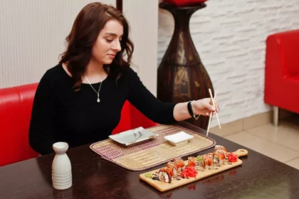Mulher sentada em mesa de restaurante japonês usando hashi para pegar peça de sushi servida em tábua de madeira, com molho shoyu e saquê ao lado