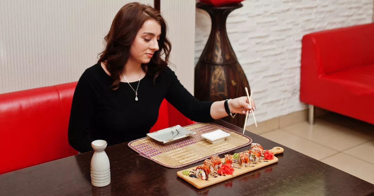 Mulher sentada em mesa de restaurante japonês usando hashi para pegar peça de sushi servida em tábua de madeira, com molho shoyu e saquê ao lado