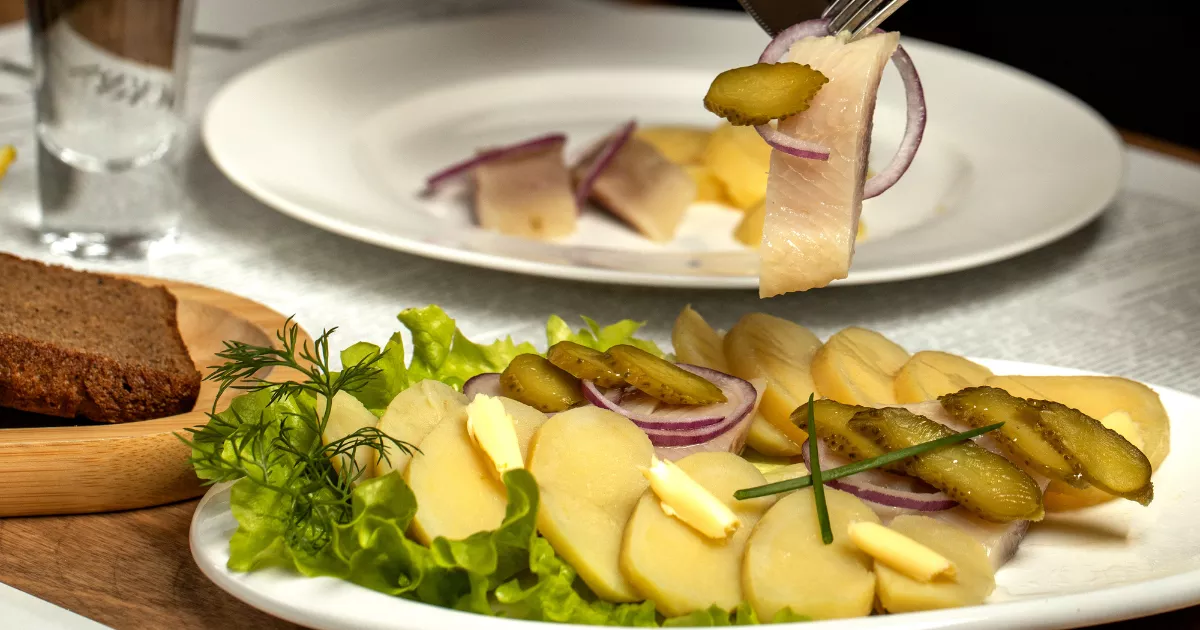 Prato executivo com batata cozida, pepino em conserva, cebola roxa e alface, com garfo erguendo fatia de peixe branco, pão escuro e endro ao fundo