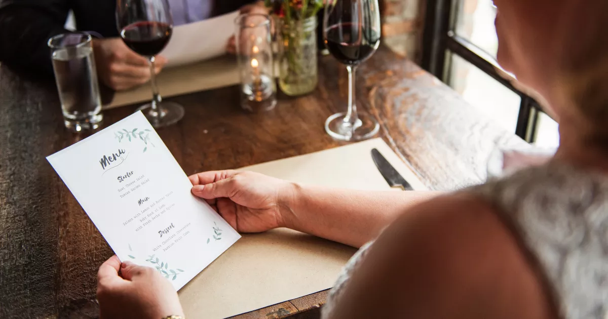 Cliente segurando cardápio branco com seções de entrada, prato principal e sobremesa em mesa de restaurante com taças de vinho e vela ao fundo