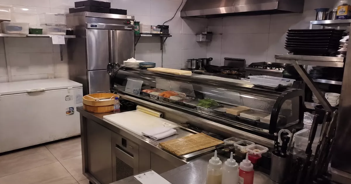 Cozinha de restaurante japonês com balcão refrigerado, peixes expostos, hangiri de madeira, makisu sobre a bancada, freezer horizontal e fogão industrial ao fundo