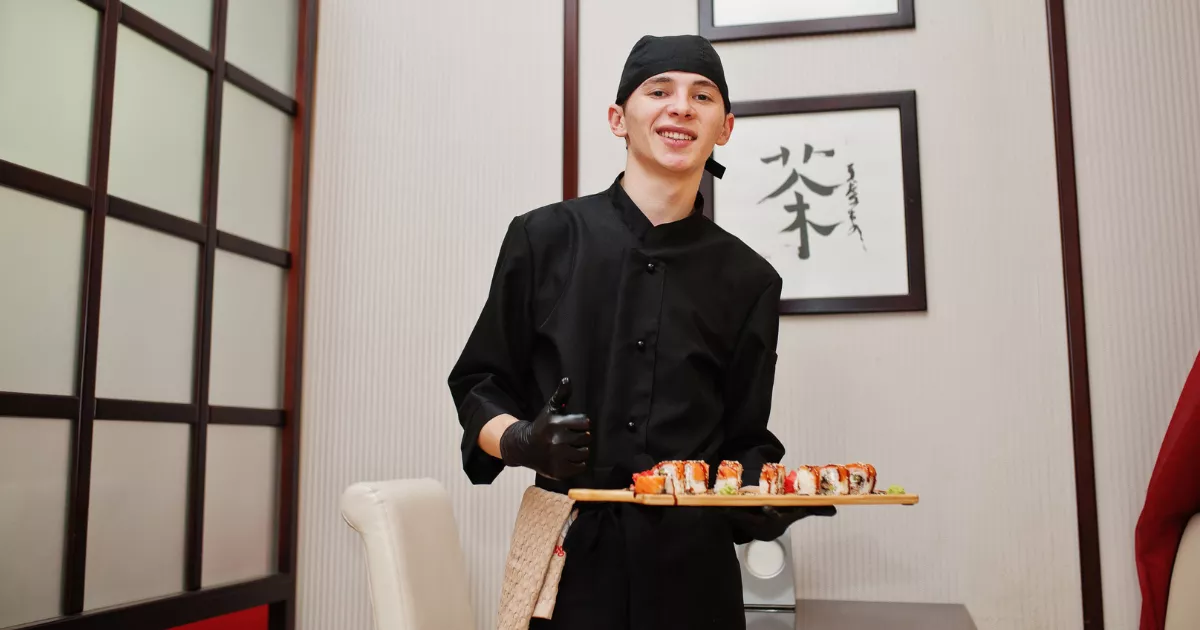 Sushiman jovem de uniforme e touca pretos sorrindo e segurando tábua com peças de sushi, com painel shoji e quadros de caligrafia japonesa ao fundo