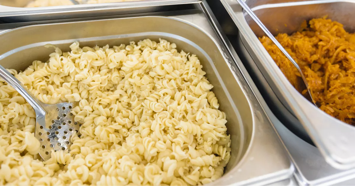 Cubas gastronômicas de inox em buffet térmico com macarrão parafuso e guarnição alaranjada, pegador de servir apoiado na cuba, em restaurante self service