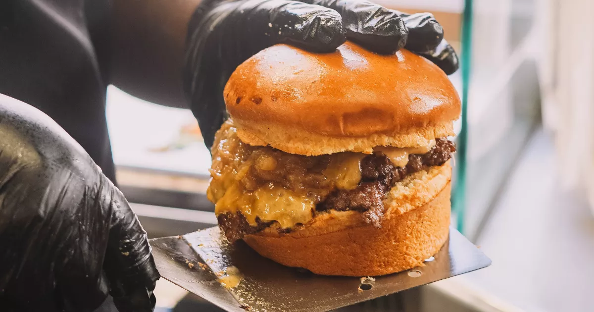 Mãos com luvas pretas segurando hambúrguer smash com queijo cheddar derretido sobre espátula de inox, pronto para embalar em cozinha de hamburgueria delivery