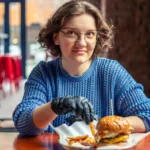 Jovem de óculos e suéter azul sentada à mesa de hamburgueria com combo de hambúrguer e batata frita, banquetas vermelhas e balcão ao fundo