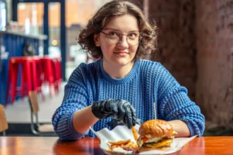 Jovem de óculos e suéter azul sentada à mesa de hamburgueria com combo de hambúrguer e batata frita, banquetas vermelhas e balcão ao fundo
