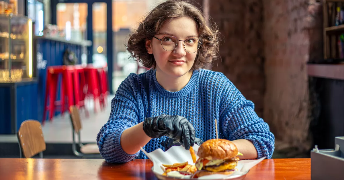 Jovem de óculos e suéter azul sentada à mesa de hamburgueria com combo de hambúrguer e batata frita, banquetas vermelhas e balcão ao fundo