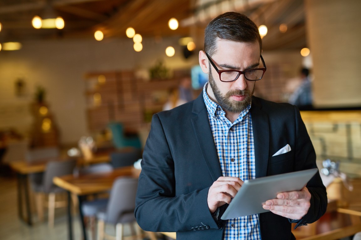 Retrato da cintura para cima de um gerente barbudo concentrado, de óculos, verificando e-mails comerciais em um tablet digital enquanto está em um restaurante moderno e espaçoso