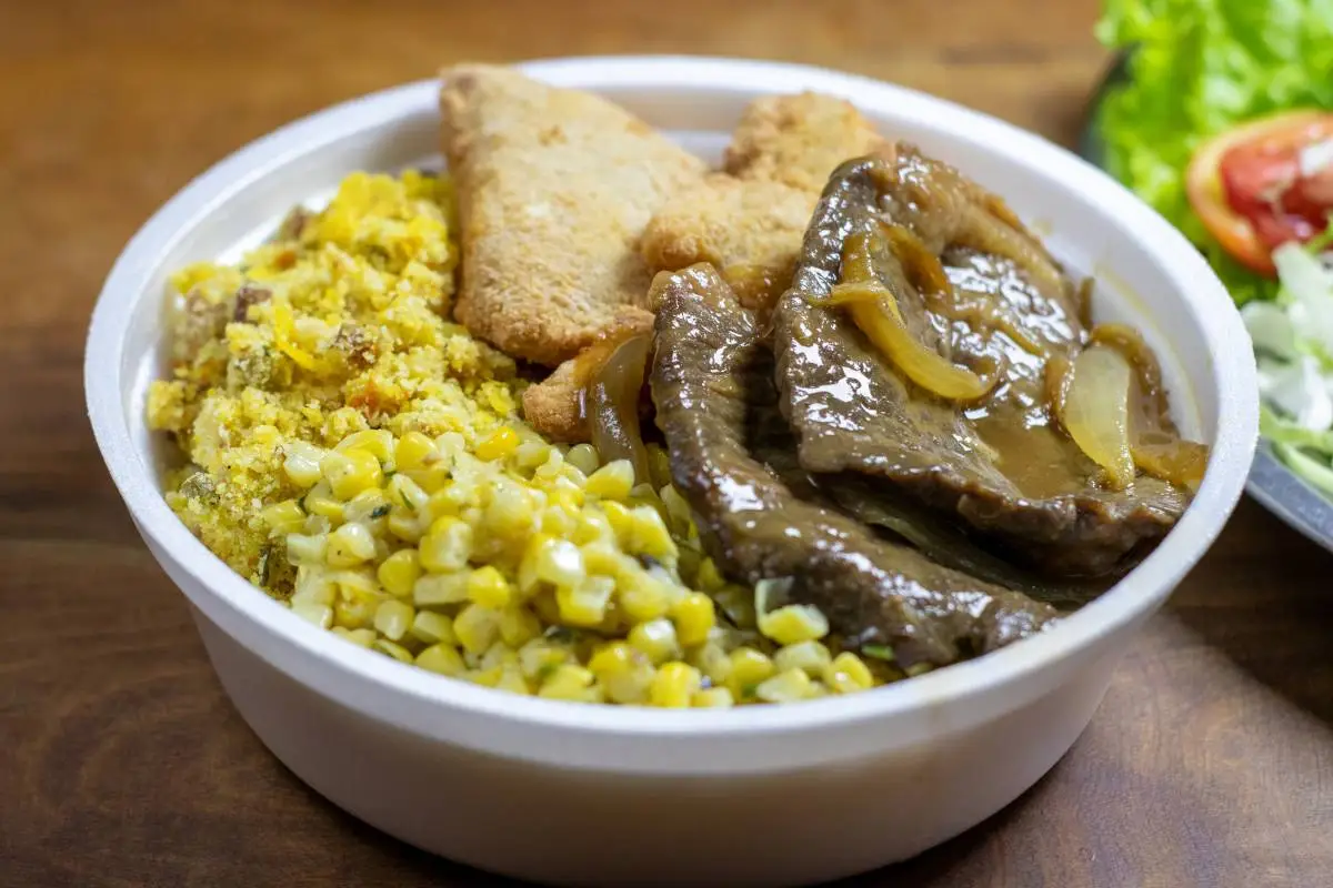 Marmita com carne, frango empanado, milho e farofa em embalagem descartável.