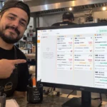 Dono de hamburgueria brasileira aponta para monitor que exibe a Central de Delivery do ControleNaMão, com cozinha e balcão ao fundo.