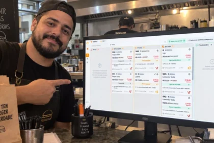 Dono de hamburgueria brasileira aponta para monitor que exibe a Central de Delivery do ControleNaMão, com cozinha e balcão ao fundo.