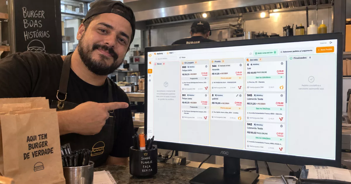 Dono de hamburgueria brasileira aponta para monitor que exibe a Central de Delivery do ControleNaMão, com cozinha e balcão ao fundo.