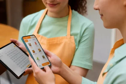 Duas atendentes de hamburgueria com aventais laranja usam celular e tablet com telas do ControleNaMão para gerenciar pedidos online.