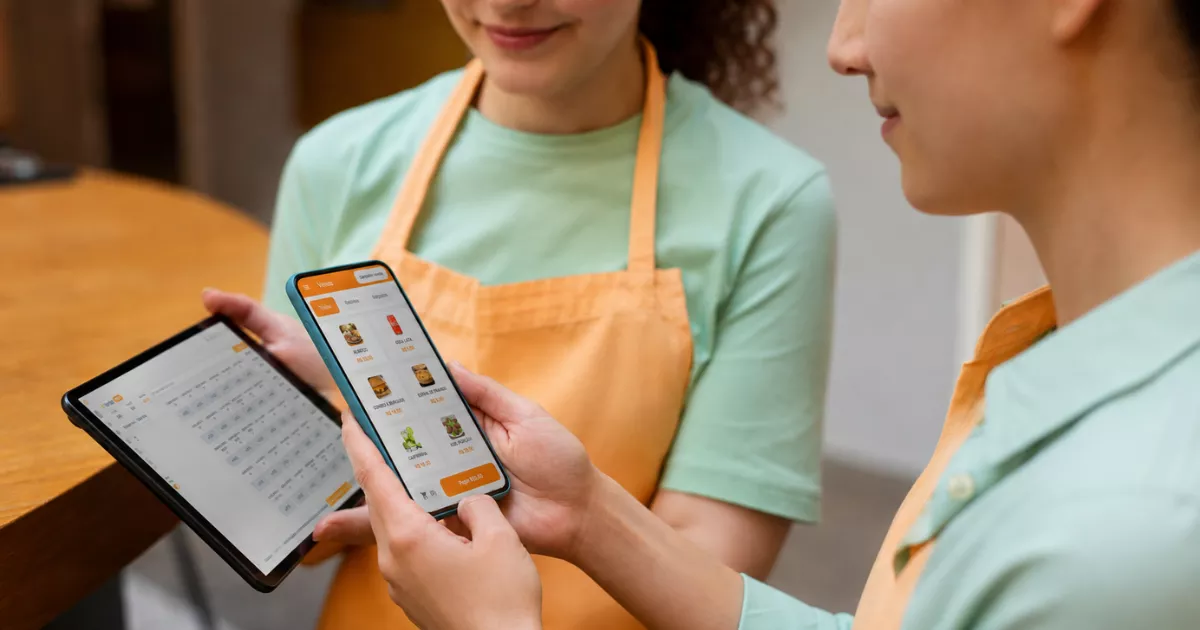 Duas atendentes de hamburgueria com aventais laranja usam celular e tablet com telas do ControleNaMão para gerenciar pedidos online.