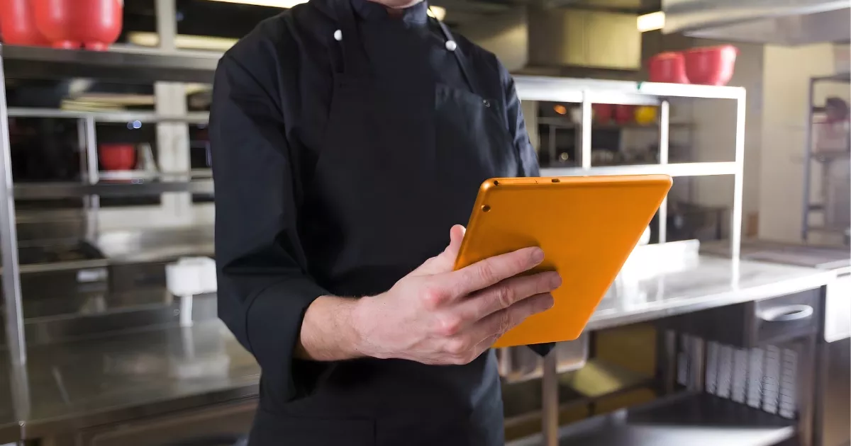 Cozinheiro de uniforme preto consultando tablet laranja em cozinha profissional com prateleiras de inox e panelas vermelhas ao fundo