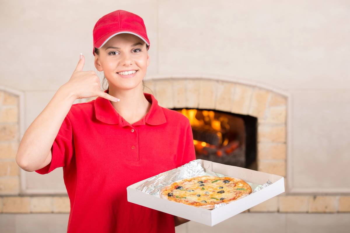 Entregadora de pizza sorridente, com uniforme vermelho, fazendo gesto de "ligação" com a mão e segurando uma caixa de pizza recém-saída do forno. Ideal para ilustrar o serviço de delivery rápido e simpático.