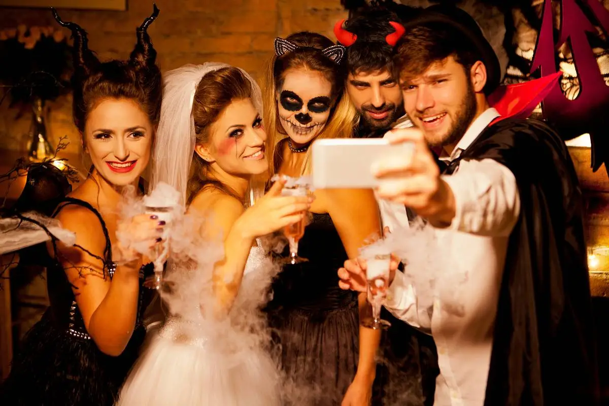 Grupo de amigos fantasiados para o Halloween tirando selfie com bebidas temáticas e fumaça decorativa.