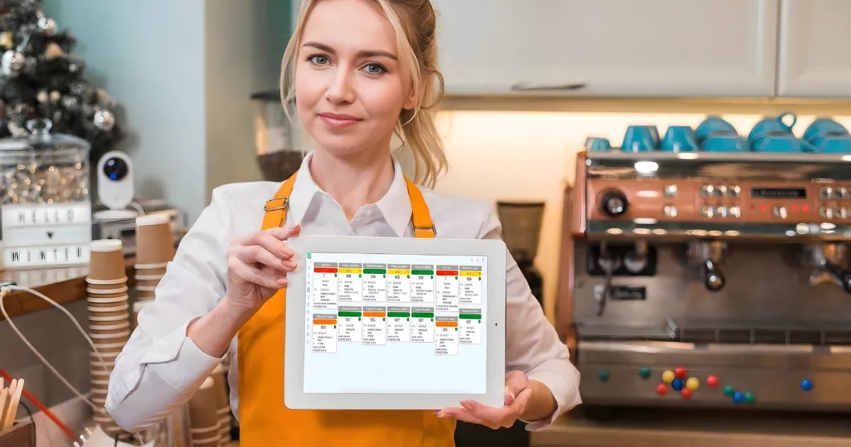 Atendente de avental laranja segurando tablet com tela do sistema KDS exibindo pedidos organizados por cards coloridos, cozinha de cafeteria ao fundo com máquina de espresso