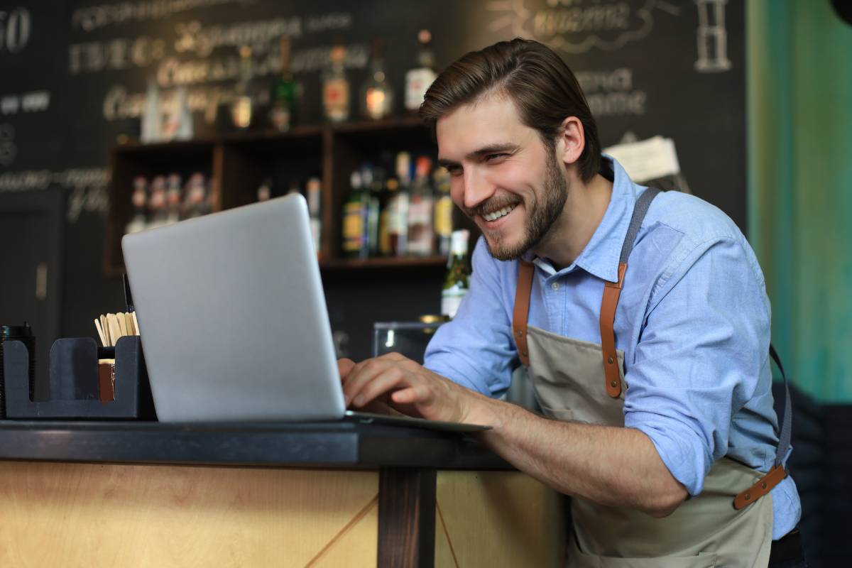 Gerente de bar sorrindo enquanto trabalha em seu laptop no balcão do restaurante.