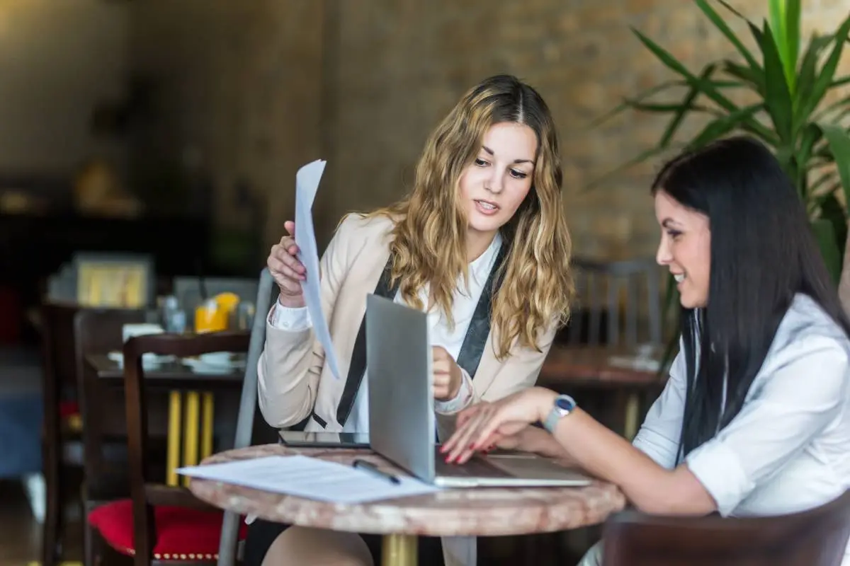 Duas mulheres discutindo estratégias de gestão em um restaurante, utilizando um laptop e documentos para otimizar a operação do negócio