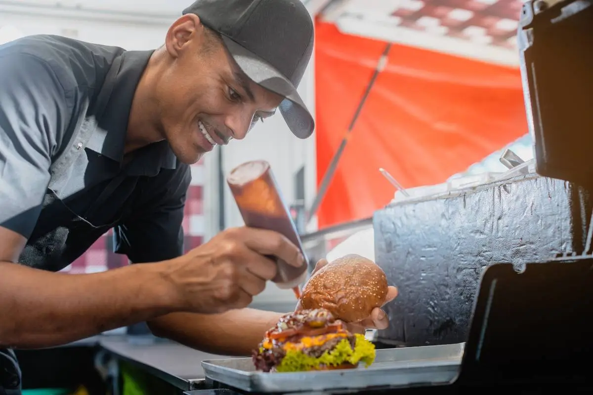 Homem preparando hambúrguer em uma hamburgueria, adicionando molho ao lanche com atenção aos detalhes.