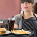 Atendente segura bandeja com hambúrguer, batatas fritas e refrigerante no salão de uma hamburgueria pequena.