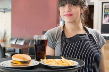 Atendente segura bandeja com hambúrguer, batatas fritas e refrigerante no salão de uma hamburgueria pequena.