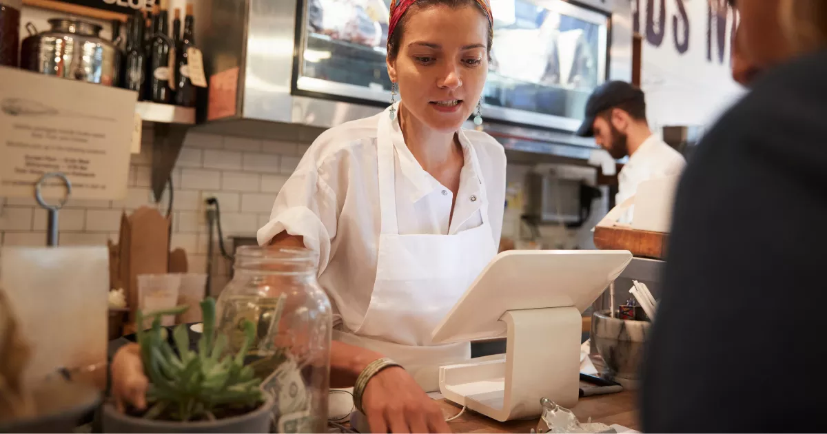 Atendente de hamburgueria opera tablet no caixa diante de cliente, com cozinha e outro funcionário trabalhando ao fundo.