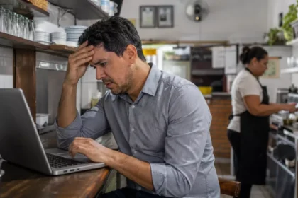 Gestor preocupado consulta um notebook no balcão enquanto uma funcionária trabalha ao fundo da cozinha de um restaurante.