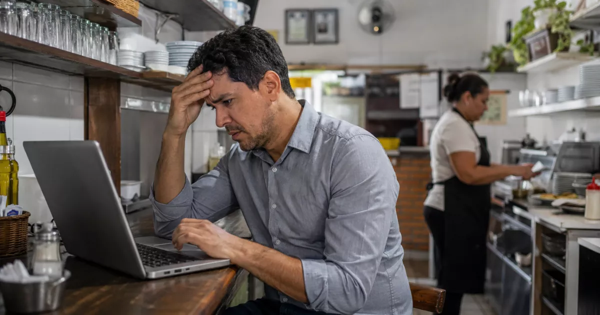 Gestor preocupado consulta um notebook no balcão enquanto uma funcionária trabalha ao fundo da cozinha de um restaurante.