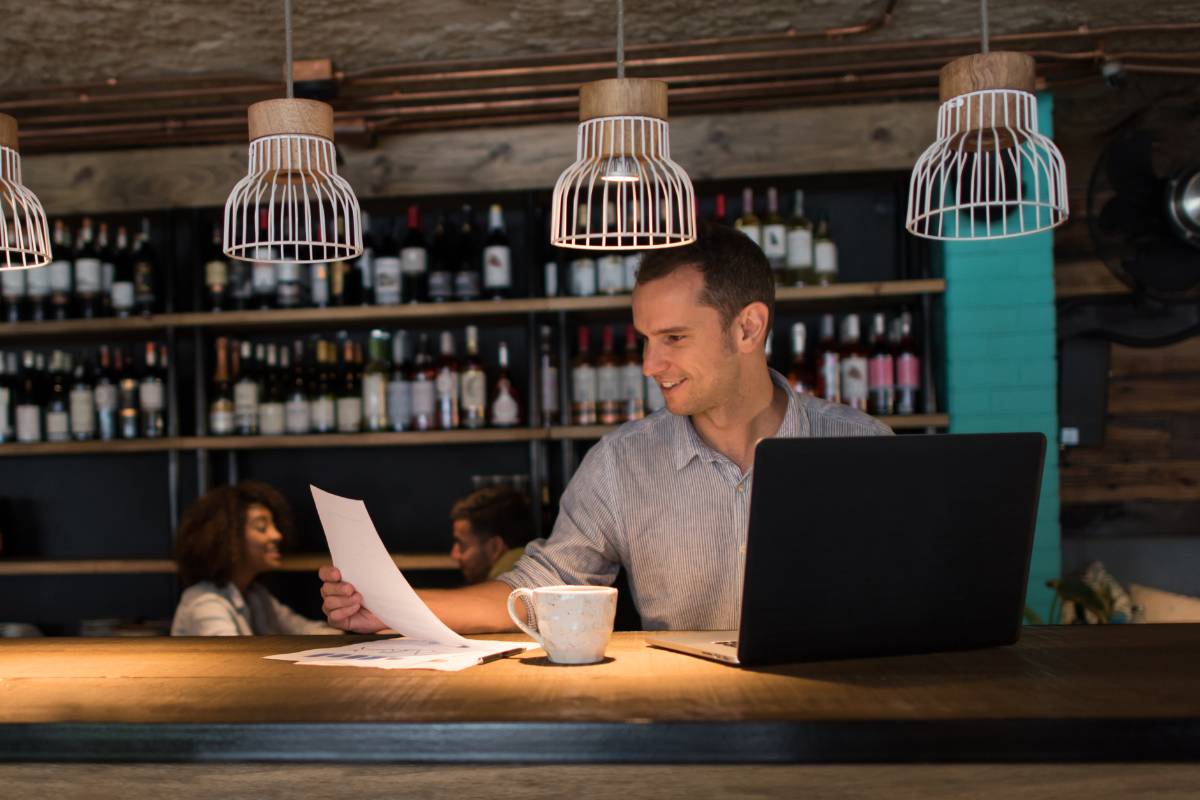 Gerente de restaurante analisando relatórios financeiros no balcão, com um laptop e uma xícara de café ao lado, em um ambiente acolhedor de bar.