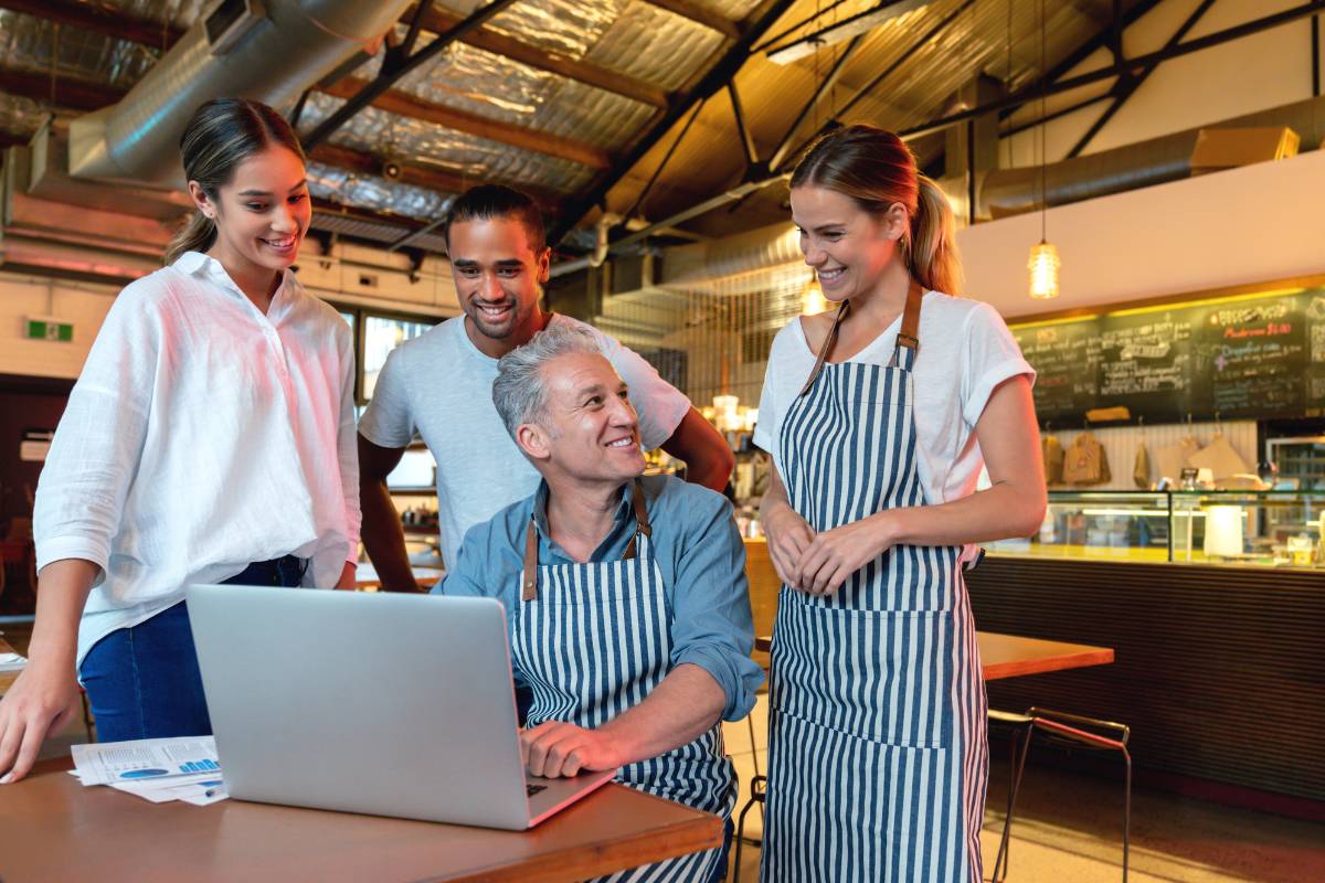 Equipe de restaurante reunida em volta de laptop com gerente, discutindo dados e estratégias de negócios, simbolizando trabalho em equipe e administração digital.