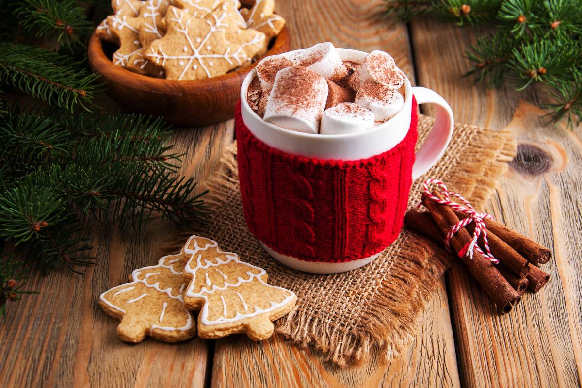 Caneca decorada com malha vermelha contendo chocolate quente com marshmallows, ao lado de biscoitos e enfeites natalinos.